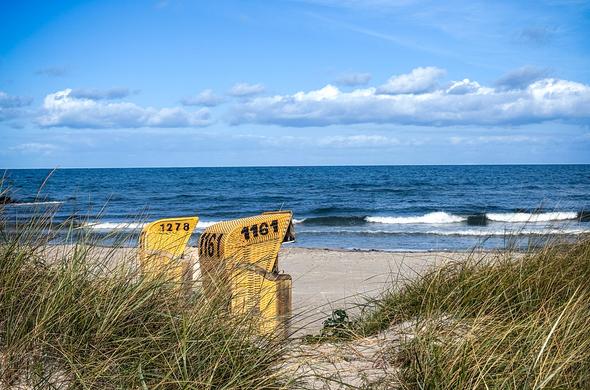Blick auf die Ostsee. Im Vordergrund Dünengräser mit schmalem Sandweg in der Mitte. Links hinter den Gräsern stehen zwei braune Strandkörbe, die man von der Seite sieht. Hinter dem feinen Sandstrand ein paar kippende Wellen in der tiefblauen Ostsee. 
Am ansonsten blauen Himmel mittig ein Wolkenband.