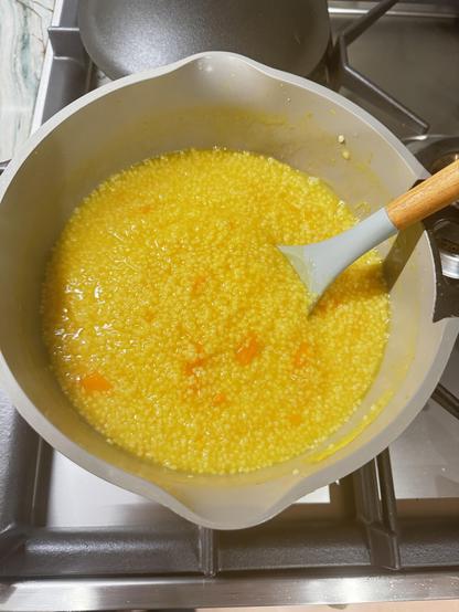 Color photo that is a close-up of a pot on a stove. Inside the pot is bright yellow cooked millet with little pieces of orange squash. A spoon with a wooden handle is sitting in the pot