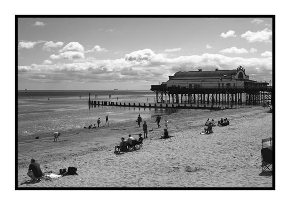 A black-and-white photograph of Cleethorpes Pier on a bright summer’s day. The pier stretches out over the sea on tall supports, its roofline and “Fish & Chips” sign clearly visible. In the foreground, people are scattered across the sandy beach: some sit in deck chairs, others walk along the waterline, while children play near the groynes leading out into the shallows. Above, a sky of billowing clouds hangs over the calm horizon.