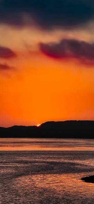 The image depicts a serene sunset over a body of water, likely a river or lake. The sky is a gradient of warm colors, transitioning from deep orange near the horizon to darker shades of blue and purple higher up. The sun is partially visible, just above the silhouette of a distant hill or mountain range, casting a warm glow on the clouds and the water's surface. The water reflects the vibrant colors of the sky, creating a shimmering effect. The overall scene is tranquil and picturesque, with the interplay of light and shadow adding depth to the composition.