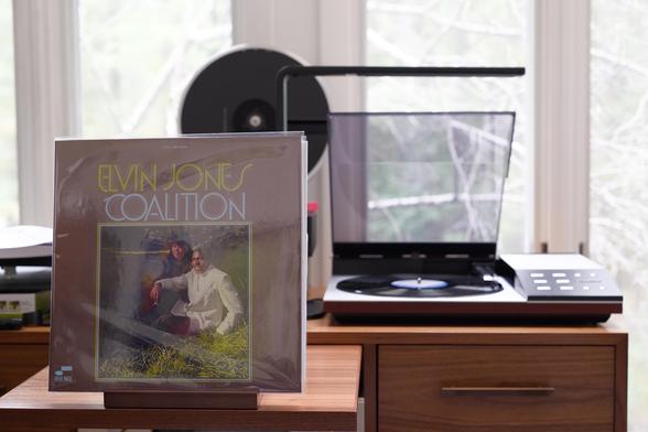 Elvin Jones - Coalition LP cover.

A very 70s looking cover, with a color picture of Elvin and his wife sitting on grass, centered at the bottom, and the artist and title above.

The black LP plays on a vintage, restored Bang & Olufsen Beogram 8002 linear-tracking turntable to the right.