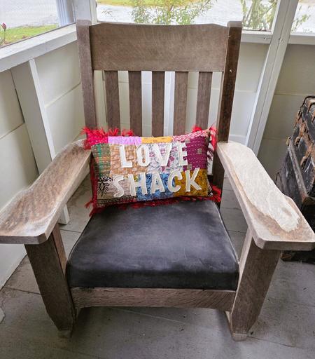 A rustic wooden armchair with wide, flat arms sits on a gray-painted porch floor. The seat cushion is black and worn smooth with age. Resting across the chair’s back is a colorful patchwork pillow stitched with bold letters that read “LOVE SHACK.” The pillow has red fringe trim and bright fabric squares in pink, orange, teal, mustard, and white. The chair is placed against light-colored porch walls with screened windows looking out toward a gravel driveway and greenery.