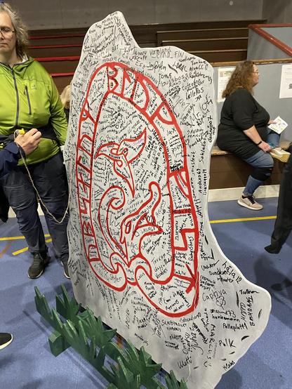 The "log book" (visitor log) for the Block Party event. It's a big wooden cutout (almost as tall as the person standing in the picture) shaped and painted like a Norse runestone. People have used markers to write their names all over it.