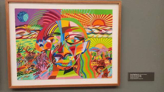 This artwork, Nuclear Pantheon #1, is by Frank Big Bear, Jr., an Ojibwe artist. Created in 1995, the piece is a vibrant and complex composition dominated by a large, fragmented face at its center. The face is rendered in a kaleidoscope of bold, contrasting colors—bright greens, yellows, reds, and blues—and appears to be split down the middle, with each side featuring different patterns and designs.
​The face is set against a dynamic background that also seems to be divided. The left side depicts a watery, marsh-like scene with a blue-and-purple moon above. The right side shows a desert landscape with a radiating sun, and various stylized plants and animals are interwoven throughout the composition. The entire piece is bursting with energy and detail, creating a powerful blend of abstracted and representational imagery.