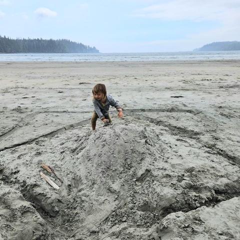 Young child playing on a sandy beach, climbing on top of a large sand pile with a serene ocean and forested horizon in the background.