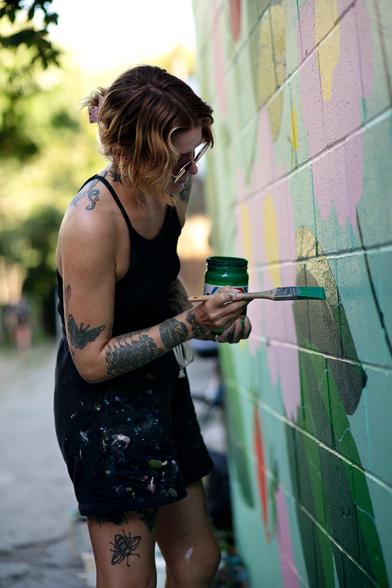 A streetart female artist painting a mural with a brush. The wall is green and pink and a warm yellow-green light is surrounding her.