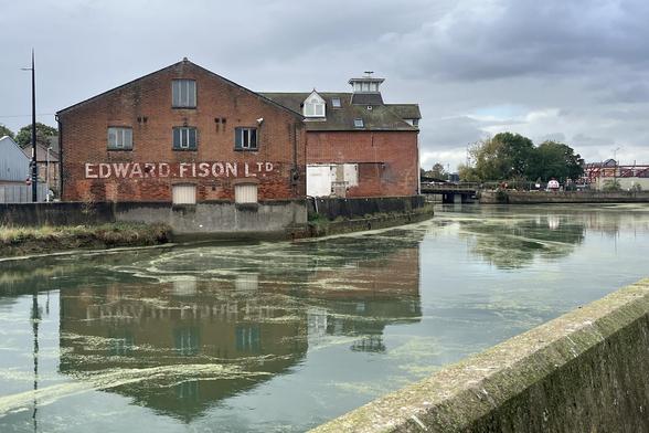 Photo across a stretch of water showing a red brick industrial building of three floor with a peaked roof. Between the ground and first floor is a horizontal band of simple white painted block lettering that reads “Edward Fison Ltd”. The entire structure and the lettering are reflected in the water.