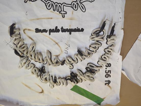 Two neon tubes bent in script to form a moon shape on a paper template pattern. The electrodes are already on and the tubes are pumped and dipped in paint. Some green tape holds the paper together.