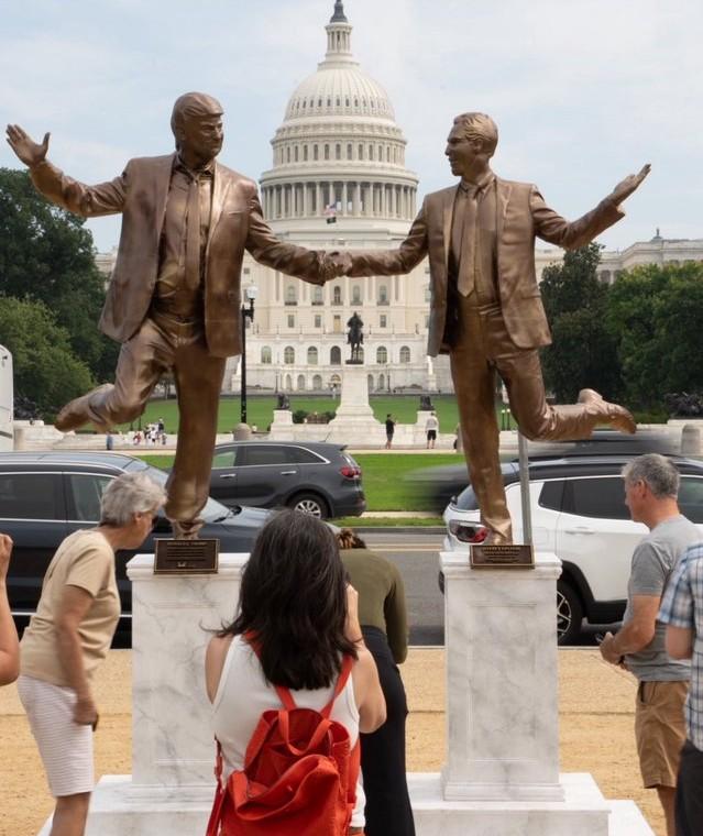 Satirical bronze-colored statues of Donald Trump and Jeffrey Epstein on the National Mall in Washington, D.C., posed holding hands with one leg raised each, in front of the U.S. Capitol building. Several people stop to look and take photos of the installation.