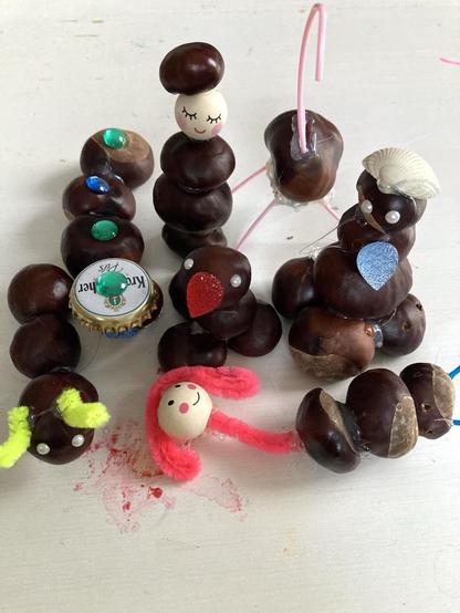 Kastanienfiguren. Links eine Raupe aus drei Kastanien mit gelben Fühlern, dann eine Raupe aus vier Kastanien, die einen Kronenkorken als Hut trägt mit, der mit einem grünen Stein (Aufklebesteine als Sticker) verziert ist, auch die anderen Kastanien der Raupe haben n der Mitte oben so einen Stein, wobei ein blauer dabei ist. Die nächste Figur hinten steht aufrecht. Drei Kastanien sind zusammengeklebt (Klebepistole), darauf befindet sich eine helle Holzkugel mit Frauengesicht, geschlossene Augen, lange Wimpern, Lächeln, rosa Backen. Vor ihr sitzt eine Vogelfigur. Roter Schnabel (auch Aufklebesticker Tropfenform) zwei kleine Perlenaugen. Davor liegt eine menschliche Figur aus drei Kastanien, langem roten Spielpfeifenreinigerhals, einem der hellen Holzkugelköpfe, dieser hat aber offene Augen und ein breites Lächeln. Sie trägt lange Haare ebenfalls aus Pfeifenreiniger Leuchtrot, die Beine sind blau, ganz dünn und nur ein klein wenig zu sehen. Es gibt noch zwei weitere Figuren auf dem Foto. Der Untergrund ist weiß.