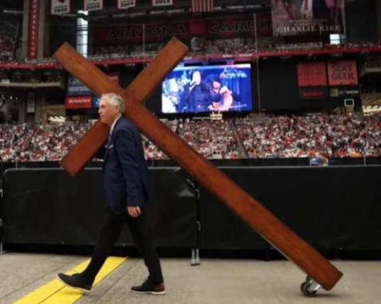 Photographie d'un homme blanc en costume-cravate portant au centre d'un stade rempli à craquer une grande croix posée sur des roulettes