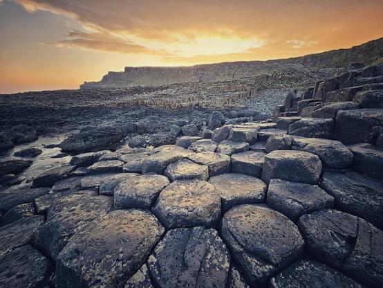 A wide-angle, low-view shot of the Giant's Causeway at sunrise. Hexagonal basalt columns fill the foreground, with smaller, broken columns receding into the distance towards a large cliff face. The sky is a blend of soft orange and yellow hues, with a few clouds scattered across the horizon. The overall mood is serene and dramatic.
