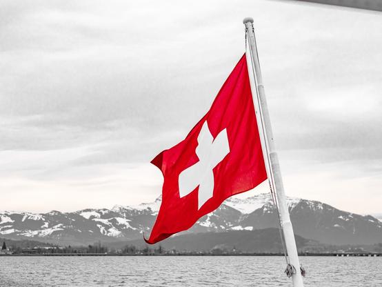 The red flag with the white cross waves at the back of a boat. The rest of the image with the lake and the mountains in the background is black and white except for the red in the flag