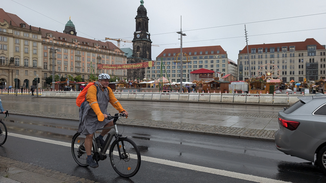OB Hilbert ist auf einem silbernen E-Bike zu sehen. Er trägt einen Regenponche, kurze Hosen und einen Helm. Im Hintergrund ist der Altmarkt wo gerade der Herbstmarkt stattfindet.