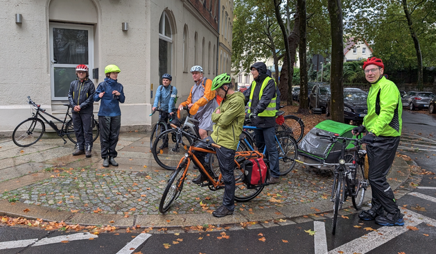 Die Gruppe ist versammelt an einer Straßenecke mit markierter Sperrfläche. Alle tragen Regenkleidung. Herr Hilbert hört den Ausführungen einer Frau zu.