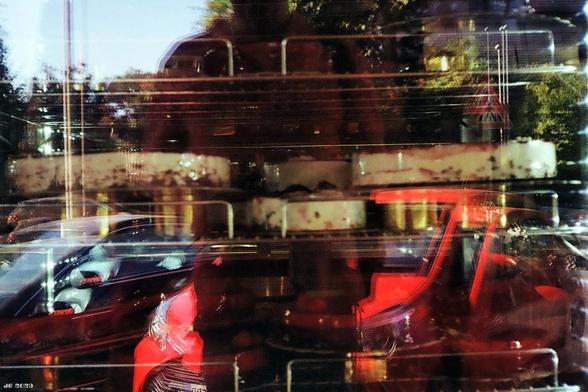 Farbfoto - Blick in eine Scheibe, in der sich verschiedene Ebenen spiegeln, darunter geparkte Autos, Gebäude, ein Metallgestell, Bäume und ein Stück Himmel. Konica C35 auf Lomo Color Negative 100.