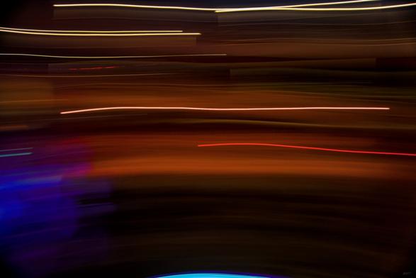 Das Foto zeigt abstrakte Lichtspuren vor einem dunklen Hintergrund, die von oben nach unten leicht bogenförmig verlaufen. Die Farbtöne reichen von warmen Orangetönen bis zu einzelnen roten und violetten Linien, im unteren Bereich sind auch bläuliche Flächen sichtbar. Das Bild ist eine Langzeitbelichtung mit Bewegungsunschärfe, wodurch die Lichtquellen zu Streifen verzogen sind und eine dynamische, fast malerische Wirkung entsteht. Es lassen sich keine klar erkennbaren Objekte oder Personen ausmachen, stattdessen steht die Bewegung und Farbstimmung im Vordergrund.
