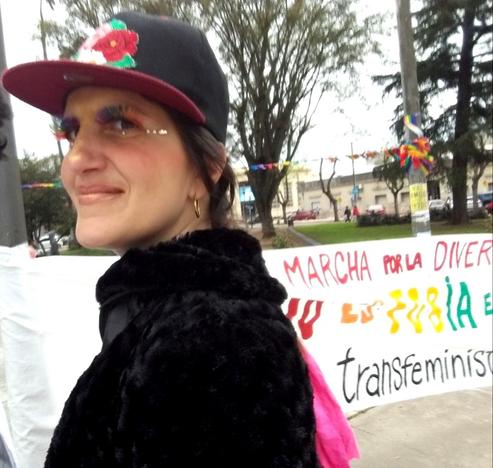 Una imagen de una plaza, al fondo se ve un cartel de letras de colores y en primer plano la imagen de Anael con maquillaje, brillos, el pelo trenzado y una gorra con visera y un detalle floral