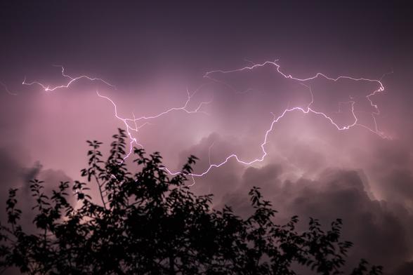 Das Bild zeigt eine dramatische nächtliche Szene mit Gewitterblitzen am Himmel. Mehrere helle Blitze ziehen sich verzweigt über den Himmel, ihre Lichtspuren wirken fast wie Adern, die sich durch die Wolken schlängeln. Der Himmel selbst ist in ein bläuliches Licht getaucht, was die Szenerie sehr eindrucksvoll und fast mystisch erscheinen lässt. Im unteren Teil des Bildes erkennt man die dunkle Silhouette von Bäumen, deren Umrisse scharf gegen das grell erleuchtete Gewitter leuchten. Die Wolken sind mächtig und bedrohlich, teilweise von den Blitzen durchdrungen.