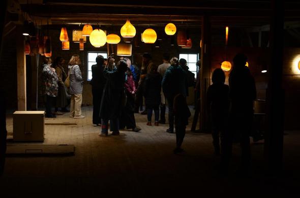 Un groupe d'adultes et enfants sont dans un grenier, éclairé par une quinzaines de lampe à abat-jours en bois accrochées au plafond diffusant une lumière chaude.