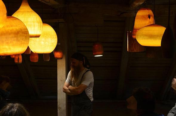 Dans un local en sous-pente, un homme barbu, un foulard cachant ses cheveux, se tient souriant les bras croisés entre deux rangées de lampes en bois.