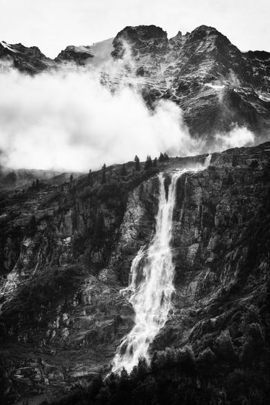 Ein majestätischer Wasserfall inmitten einer bergigen Landschaft. Der Wasserfall stürzt von einer steilen Felswand hinab und schlängelt sich durch das felsige Terrain. Die umliegenden Berge sind schroff und mit spärlicher Vegetation bedeckt. Über den Gipfeln hängen dichte Wolken, die sich mit dem Nebel vermischen und eine geheimnisvolle Stimmung erzeugen. Die Szene ist in Schwarz-Weiss gehalten, was die Kontraste zwischen den dunklen Felsen, dem hellen Wasser und den weichen Wolken besonders betont. Die Atmosphäre wirkt wild, kraftvoll und gleichzeitig friedlich.