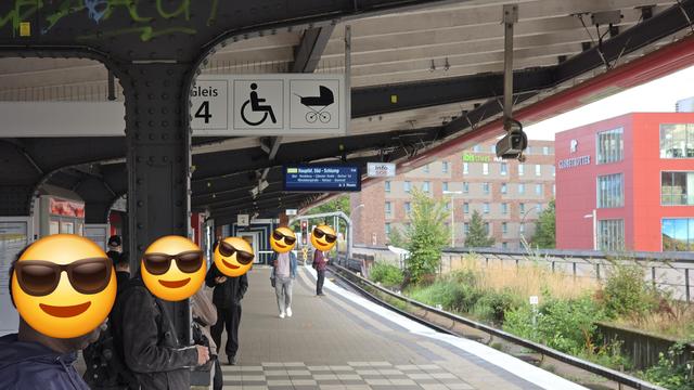 Es ist ein Bahnsteig mit neuer Fahrtanzeigetafel mit mehreren Personen mit Smiley zur Zensierung der Gesichter zu sehen.  Im Hintergrund sieht man zwei hohe Gebäude.