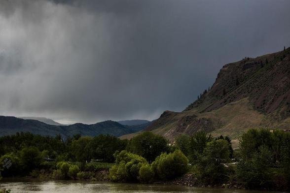 Looking North across the South Thompson River. Lush vegetation on the other bank, merging into dry grasslands. In the middle distance a rocky outcrop with scattered trees. In the far distance, mountains and mesas, with fingers of rain reaching down from a cloudy sky.