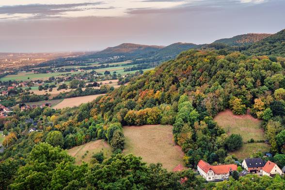Die hochauflösende Aufnahme zeigt eine malerische Landschaftsansicht im Übergang vom Spätsommer zum Frühherbst. Das Bild wurde von einem erhöhten Standpunkt aus aufgenommen, vermutlich einem Hügel, und blickt über eine weite, sanft abfallende Landschaft.
Im Vordergrund dominieren dichte, bewaldete Hügel, die in einer Mischung aus grünen, gelben und orangefarbenen Farbtönen leuchten. Einzelne Lichtungen unterbrechen den Wald und zeigen trockenes, goldbraunes Gras. Am unteren rechten Bildrand sind einige traditionelle, rot gedeckte Fachwerkhäuser zu sehen, die in die Landschaft eingebettet sind.
Die Mitte des Bildes wird von einem weitläufigen Tal eingenommen, das durch landwirtschaftliche Felder und kleine Dörfer strukturiert wird. Die Felder variieren in der Farbe, von tiefem Grün bis zu hellbraun und Gelb. In der Ferne, am linken Horizont, ist eine große Stadt sichtbar, die mit ihren Fabriken und Industrieanlagen einen starken Kontrast zur ländlichen Idylle im Vordergrund bildet.
Der Himmel ist klar und weit, mit sanften Wolkenformationen. Das späte Morgenlicht taucht die Szene in ein warmes, weiches Licht, das besonders die Hügel und die Baumkronen zum Leuchten bringt. Die Atmosphäre wirkt ruhig und friedlich.