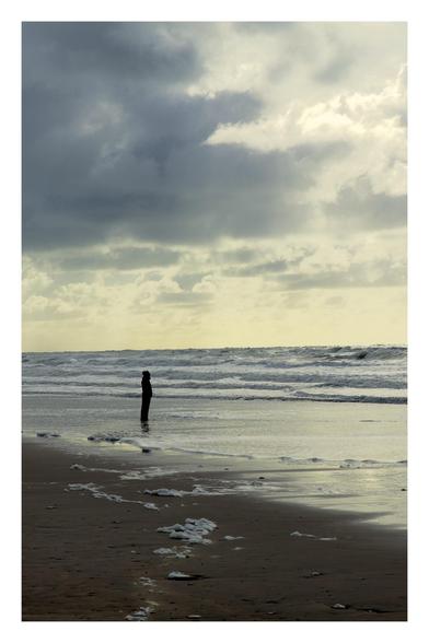 Eine einzelne Person steht mit den Füßen in einem stürmischem Meer. Die Person schaut in Regenjacke in die Ferne. Im Himmel sind dunkle Wolkenformationen zu sehen.