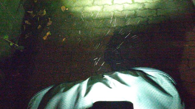 Eine Fahrradfronttasche im Licht eines am Lenker befestigten Fahrradscheiwerfer. Die Tropfen des vom Wind verwirbelten Nieselregens erscheinen als dünne Striche in verschiedene Richtungen. Das Bild hat vor allem kalte Grautöne und warme Grüntöne.