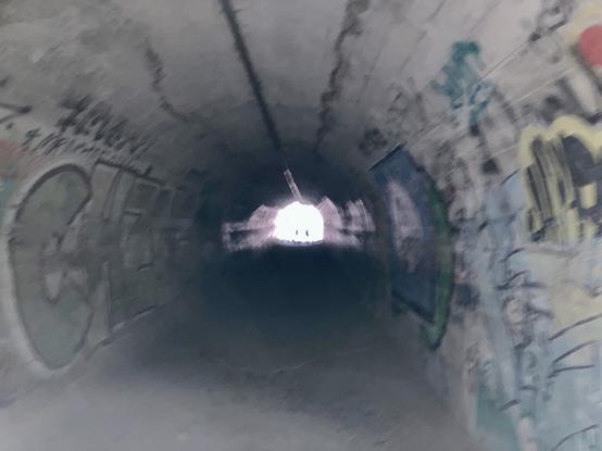 Handheld photo in dark tunnel, no flash. Light at end shows graffiti on walls