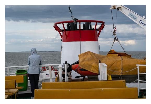 Foto im Querformat. Blick über das Deck einer Fähre. Im Vordergrund sind gelbe Sitzbänke, dahinter weiße Reling und Rettungsboote. Dahinter ein weiß-roter kleiner Turm. Links steht eine Person an der Reling und schaut über das Meer. Sie hat eine Regenjacke an, die Kapuze auf und ist mit dem Rücken zur Kamera gewandt. Der Himmel ist bewölkt.
