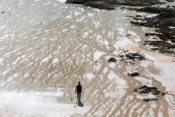 Sondengänger, der seinen Metalldetektor nach links schwenkt, am Strand von Saint-Malo. Das Wasser hat bei Ebbe viele diagonalen Furchen im Strand hinterlassen