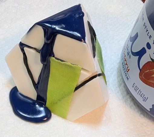 a white plastic part mould (oddly shaped: like a pyramid, but with the corners snipped off). It is held together by black silicone hair bands plus some backup green tape. The top of the mould has deep blue/indigo wax filling the top, and a trickle of it has run down the front of the mould.

Next to the mould is a glass yogourt pot that was used to melt the wax. What was clear glass on it has a thin layer of dark blue wax