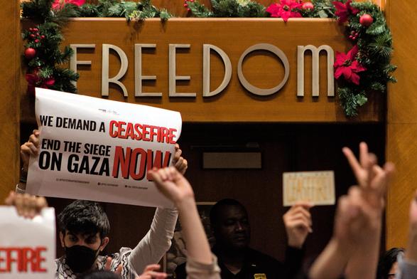 The back of the main meeting room at city hall. In frame are a few people holding up signs as well as peace signs with their hands. One sign states "We demand a ceasefire / End the siege on Gaza now" framed center-left of the image. Behind it taking up center-top portion of the image are the words "freedom". Ironic.