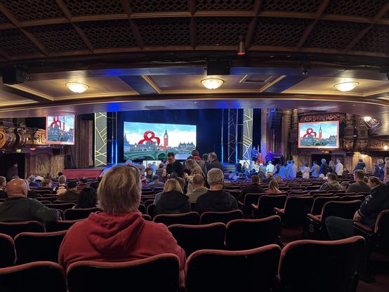 An audience gathers in a theater with ornate decor, facing a stage adorned with screens displaying images of a city skyline. Most attendees appear to be settling into their seats, while some are milling about. The theater's lighting adds a dramatic effect to the scene