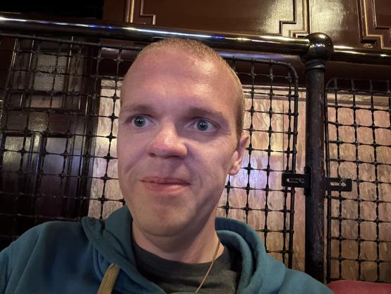 Me - Paul Douglas, a caucasian person with short hair - sitting in front of a fence inside, with a wood panelled wall behind. I am wearing a teal hoodie and smiling in anticipation. The setting has a vintage feel, with dim lighting and decorative elements.