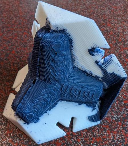a small blue four-footed structure cast roughly in wax, still surrounded by two out of four of its mould pieces. Excess wax can be seen blocking the filling port at right. There's a small holed in the object near the centre where an internal void has formed as the wax cooled and contracted. This might be as much as 10% of the total model volume