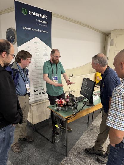 Marshall Kirk McKusick, Eric Allman, a couple of (much) younger BSD developers and myself hovering over an old open-frame PC build which is busy booting NetBSD. Sponsor pull-up poster for Entersekt/Modirum (my employer) in the background.