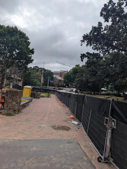 Photo of a wide brick sidewalk, separated from the street by a black construction fence. There are no signs of construction except for a stack of traffic barriers off to one side and a yellow porta-potty. In the distance is a US flag at half-staff (photo was taken on Sept 11).