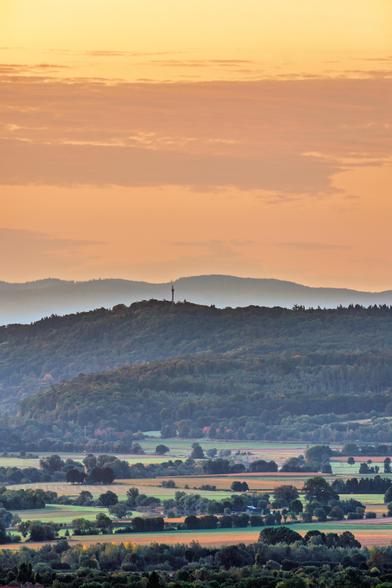 Das Bild zeigt eine weitläufige Landschaft bei Sonnenuntergang oder Sonnenaufgang. Im Vordergrund erstreckt sich ein flaches Tal mit Feldern und einzelnen Bäumen, die teils als kleine Baumgruppen oder Alleen angeordnet sind. Die Felder sind in verschiedenen Brauntönen gehalten, was auf geerntetes Land hindeutet.
Bildbeschreibung
Im mittleren Bereich des Bildes erhebt sich ein dicht bewaldeter Hügel, auf dessen Spitze ein Sendemast oder Aussichtsturm zu erkennen ist. Die Wälder auf dem Hügel sind in dunklen Blau- und Grüntönen gehalten. Hinter diesem Hügel sind weitere Hügelketten zu sehen, die in der Ferne in sanfte, bläuliche Farbtöne übergehen. Dieser Effekt, bei dem die Farben mit zunehmender Entfernung verblassen und bläulich erscheinen, wird Luftperspektive genannt.
Der Himmel über der Landschaft ist in warmen Orange-, Gelb- und Rosatönen gefärbt. Er ist mit lockeren, teils streifenförmigen Wolken bedeckt, die das weiche Licht der Sonne reflektieren. Die gesamte Komposition strahlt eine ruhige und friedliche Atmosphäre aus.