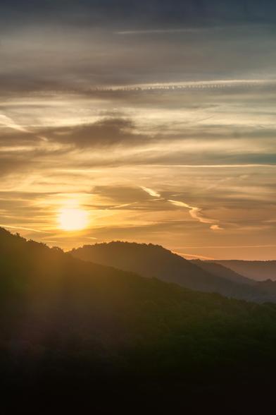 Das Bild zeigt einen Sonnenaufgang über einer Hügellandschaft. Die untergehende Sonne ist als leuchtender, weiß-gelber Lichtball am linken Rand des Bildes zu sehen und wirft einen starken Gegenlichteffekt auf die Szenerie.
Bildbeschreibung
Im Vordergrund des Bildes sind die silhouettenhaften Umrisse von mehreren bewaldeten Hügeln zu erkennen. Sie liegen in verschiedenen Ebenen, wobei der vorderste Hügel als dunkle, fast schwarze Masse erscheint. Die dahinterliegenden Hügel sind in immer helleren Blautönen dargestellt, ein Effekt, der durch die Luftperspektive entsteht.
Der Himmel dominiert das obere Drittel des Bildes und ist in warmen Orange-, Gold- und Brauntönen gehalten, die sanft in ein kühleres Blau und Grau am oberen Rand übergehen. Mehrere lange, weiße Kondensstreifen von Flugzeugen ziehen sich horizontal durch den Himmel und betonen die Weite und Höhe des Himmelsraums. Die Kombination aus den warmen Farben der untergehenden Sonne und den kühleren Farbtönen am oberen Himmelrand erzeugt einen spannungsvollen Kontrast.