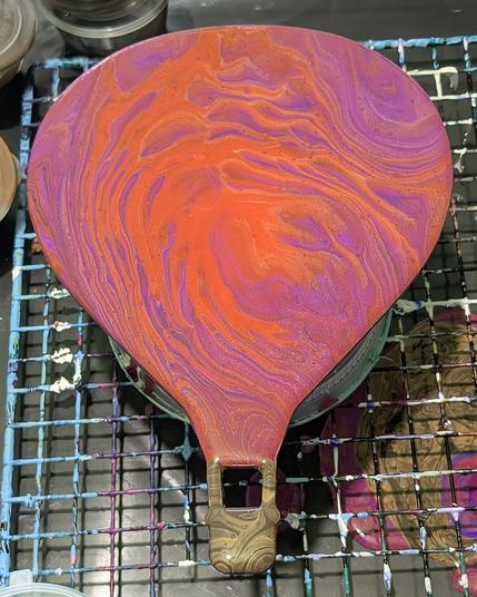 A wooden cutout of a hot air balloon painted using the ring pour fluid art technique; the main part of the balloon is orange, pink, purple, and gold while the basket is shades of brown.