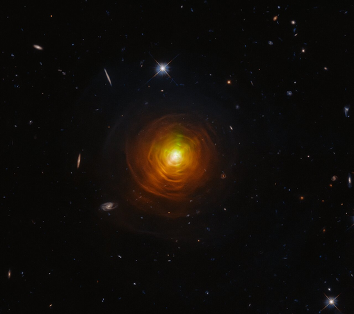 A Hubble Space Telescope image of the carbon star CW Leonis, showing a bright yellow core surrounded by swirling, glowing orange and brown dust shells. The shells form a circular, layered pattern radiating outward into the dark background of space, which is dotted with faint stars and distant galaxies.
