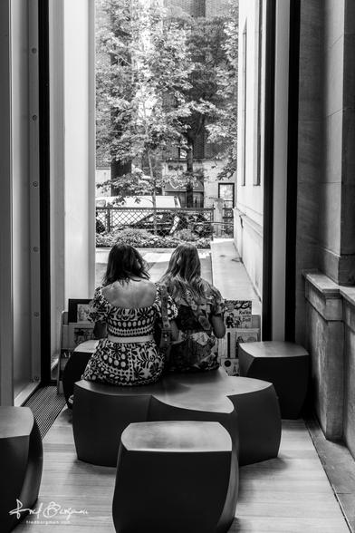 Two women sitting shoulder to shoulder on a star shaped seating area in front of a row of books and large glass window with a courtyard outside. They are inside the Morgan Library and Museum in New York City in June 2025