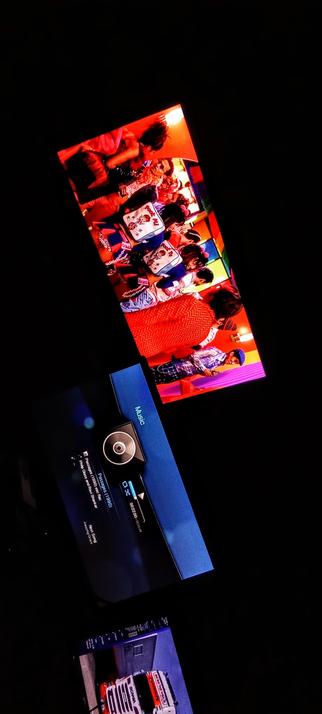 The photograph shot in total absolute darkness features a television screen displaying a vibrant scene with multiple people, likely at a social gathering or event. The scene is illuminated with warm red and orange hues, and the individuals are dressed in colorful attire, including traditional Japanese clothing. The screen is tilted at an angle, adding a dynamic perspective to the image. Below the television, there is a blue device with a circular component, possibly a music player, displaying text and a progress bar. The device is labeled "Music." The background is predominantly black, emphasizing the brightly lit screen and the device.