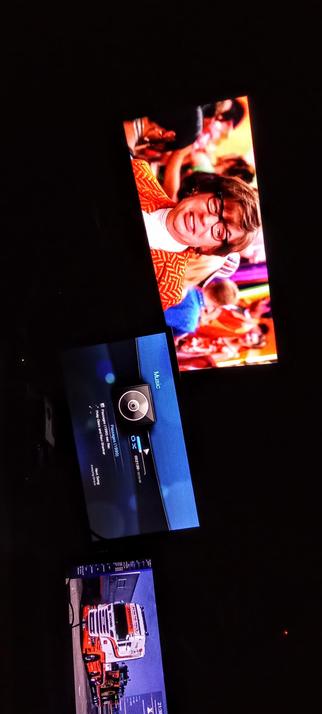 The photograph shows a dark room with three screens displaying different content. The top screen shows a person wearing glasses and a red patterned top, smiling, with a crowd in the background. The middle screen displays a music player interface with the word "Music" and a track titled "Passages (1990)" by "Philip Glass and Raw Power" by Janet Jackson. The bottom screen shows a red and white truck with the brand "Scania" on it, parked in a lot. The screens are angled and partially overlapping, with the middle screen being the most prominent.

Provided by @altbot, generated privately and locally using Ovis2-8B

🌱 Energy used: 0.574 Wh