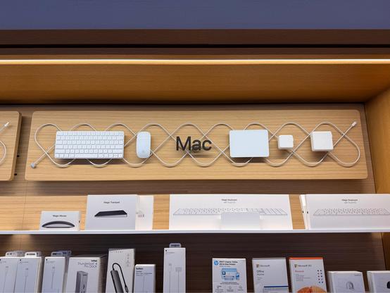 a photograph of a sign that says “mac”, surrounded by trackpad, keyboard, mouse, etc. all the actual products on stand-offs to lift them from the wooden back. there is a magsafe cable in a cool woven pattern behind them.