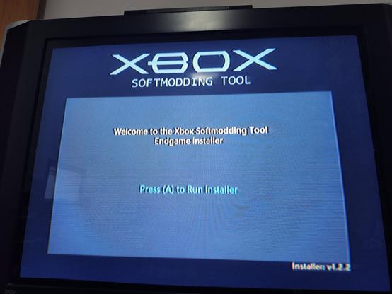Português:
A imagem mostra a tela de instalação do Xbox Softmodding Tool, exibida em uma televisão de tubo clássica. No centro da tela, aparece a mensagem de boas-vindas: “Welcome to the Xbox Softmodding Tool Endgame Installer”, convidando o usuário a pressionar o botão A do controle para iniciar o instalador. Este tipo de ferramenta é utilizada para modificar o software do Xbox original, permitindo desbloquear novas funções, instalar dashboards alternativos e executar aplicativos ou jogos de forma personalizada. O visual simples e direto traz um ar nostálgico, remetendo à época dos consoles clássicos do início dos anos 2000.

English:
The image shows the installation screen of the Xbox Softmodding Tool, displayed on a classic CRT television. In the center, a welcome message reads: “Welcome to the Xbox Softmodding Tool Endgame Installer”, instructing the user to press the A button to run the installer. This tool is used to modify the software of the original Xbox, unlocking new feature